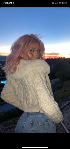 a woman with blonde hair wearing a white sweater and blue jeans standing in front of a sunset