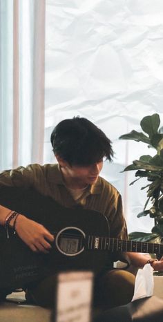 a young man sitting on the floor playing an acoustic guitar in front of a potted plant