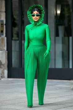 a woman in green is standing on the sidewalk wearing sunglasses and a green outfit with ruffles