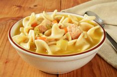 a white bowl filled with chicken noodle soup on top of a wooden table next to a spoon