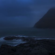 the moon is setting over the ocean with dark clouds in the sky, and waves crashing on the rocks