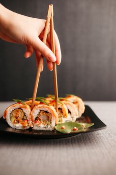 a person holding chopsticks over sushi rolls on a black plate with green leaves