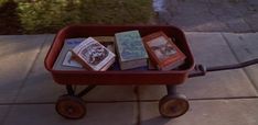 a red wagon filled with books sitting on top of a cement floor next to a sidewalk