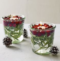 two glass cups filled with candles sitting on top of a table next to pine cones