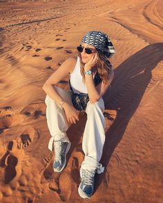 a woman sitting in the sand wearing sunglasses and a bandana on top of her head