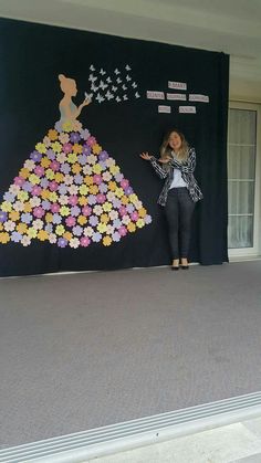 a woman standing in front of a black wall with flowers on it and an image of a dress made out of paper