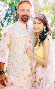 a man and woman standing next to each other in front of some trees, smiling at the camera