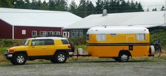 a yellow truck pulling a trailer with a camper on it's back in front of a barn