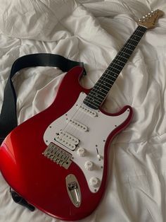 a red electric guitar laying on top of a white sheet with a black strap around it