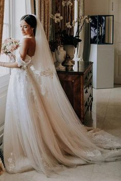 a woman standing in front of a window wearing a wedding dress and holding a bouquet