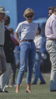 a woman in white shirt and blue jeans standing next to other people on grass field