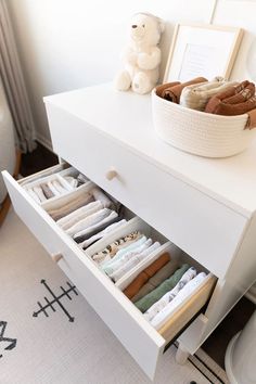 an open drawer in a white dresser next to a teddy bear and other items on the floor