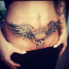 a woman's stomach with a skull and wings tattoo on her belly, which is adorned with a crown