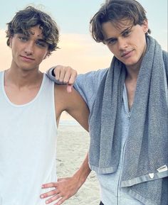 two young men standing next to each other near the ocean with towels on their shoulders