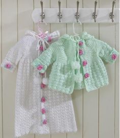 two knitted baby clothes hanging from hooks on a white wooden wall with pink and green flowers