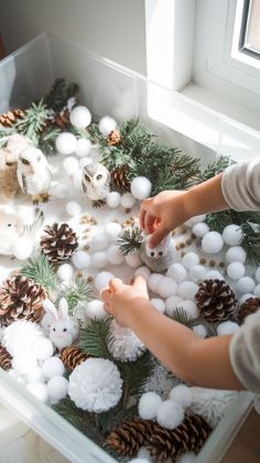 a child is playing with cotton balls and pine cones in a play box filled with snow