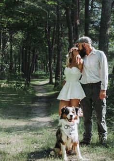 a man and woman standing next to a dog on a forest path with trees in the background