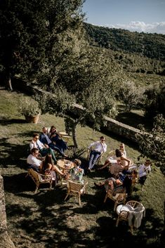 a group of people sitting on top of a grass covered field next to trees and bushes