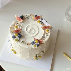 a white cake with fruit and flowers on it sitting on a table next to a pen