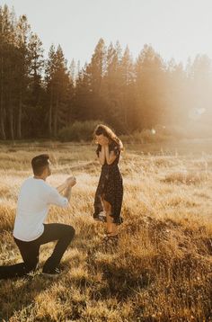 a man kneeling down next to a woman on top of a grass covered field with trees in the background