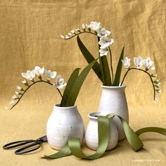 two white vases with flowers in them on a yellow cloth covered background, one is holding a green ribbon and the other has scissors