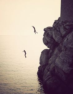 two seagulls flying over the ocean next to a rocky cliff with a lighthouse on top