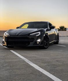 a black sports car is parked in a parking lot at sunset or dawn with the sun setting behind it