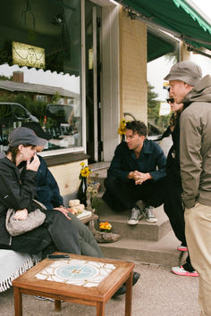 three people are sitting on the sidewalk talking on their cell phones while one person is standing