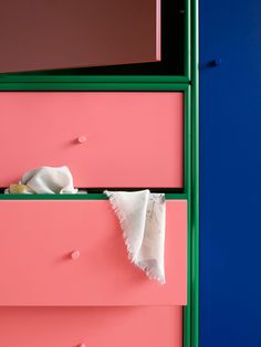 a pink and green dresser with white cloth on it's top drawer, against a blue wall