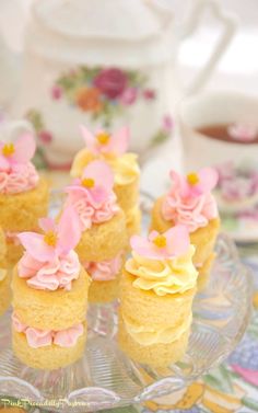 small cupcakes with pink frosting and flowers on a glass plate next to a teapot