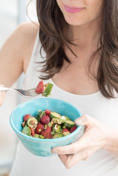 a woman holding a blue bowl with strawberries and cucumbers in it while eating