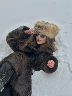 a woman laying down in the snow wearing a fur hat and sunglasses with her hands on her head