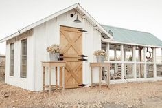 a small white shed with two planters on the outside and a table in front