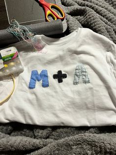 a white shirt with the letters m and m on it sitting next to scissors, thread and other crafting supplies