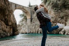 a man and woman are kissing in front of a river with a bridge behind them