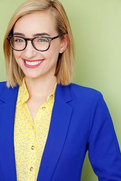 a woman wearing glasses and a blue jacket smiles at the camera while standing in front of a green wall