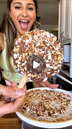 a woman holding up a giant chocolate donut with nuts on it and an ice cream sundae in front of her