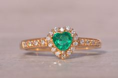 The Mademoiselle: Heart Shaped Natural Emerald and Diamond Ring in Yellow Gold. This exquisite ring features a heart brilliant-cut natural emerald, measuring 4.8mm by 4.7mm, showcasing a vivid very slightly bluish-green color. The emerald is surrounded by a halo of round brilliant-cut natural diamonds, which extend down the shank, enhancing the ring's brilliance. Expertly crafted in 14-karat yellow gold, this piece is currently a size 7.25 but can be resized to fit any finger for an additional f Gorgeous Rings, Emerald And Diamond Ring, Bluish Green, Natural Emerald, Rings Statement, Round Brilliant, A Heart, Green Color, Natural Diamonds