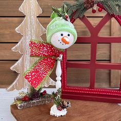 a snowman with a green hat and red bow on it's head sitting next to a christmas tree