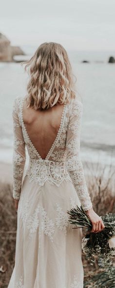 the back of a woman's wedding dress as she walks along the beach with her bouquet in hand