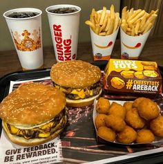 burgers and fries are sitting on a tray next to two cups of hot dogs