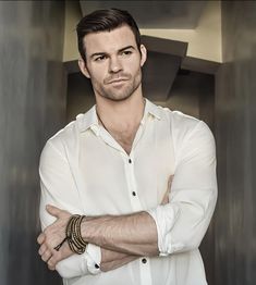 a man with his arms crossed standing in front of a gray wall wearing a white shirt