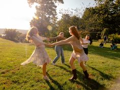 three girls in dresses are playing with each other on the grass near some trees and people