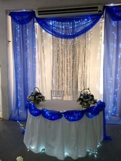 a white table topped with blue flowers under a window covered in sheer curtains and lights