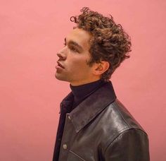 a man with curly hair wearing a leather jacket and looking off to the side against a pink background