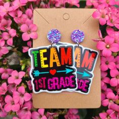 a pair of earrings with the words team and 1st grade on them in front of pink flowers