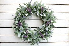 a wreath with purple flowers and greenery is hanging on a white wooden wall in front of a door