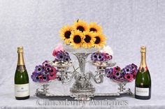 two bottles of champagne and three sunflowers on a silver tray with cupcakes