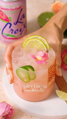 a hand holding a drink with limes and pink flowers on the rim next to a can of soda