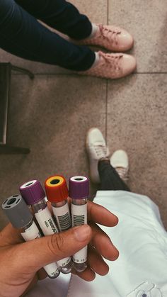 a person holding four different colored inks in their left hand and two people sitting on the floor behind them